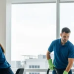 Three professionals in blue uniforms and green gloves cleaning an office—one wiping a monitor, one vacuuming, and one cleaning a window.